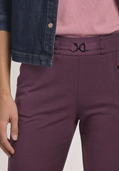 Onderlichaam van een persoon in paarse broek met decoratieve sluiting en spijkerjasje.