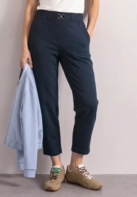 Persoon draagt marineblauwe enkelhoge broek, wit shirt, lichte sneakers en houdt een lichtblauwe jas vast.