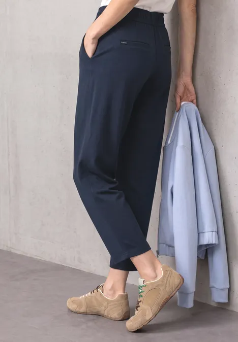Damesbeen in donkerblauwe broek, houdt lichtblauw jasje vast, draagt beige sneakers.