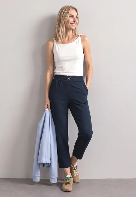 Een blonde vrouw leunt tegen de muur, draagt een wit topje en donkerblauwe broek, houdt een lichtblauwe blazer vast.