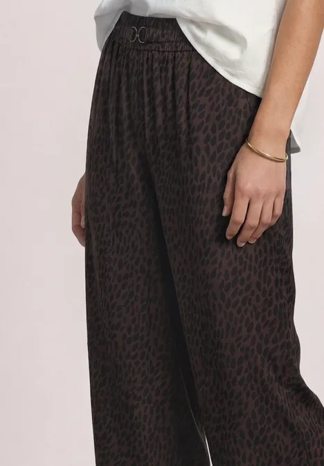 Mid Waist Straight Leg Hose mit Print molten earth Dunkelbraune, weit geschnittene Hose mit subtilem Leomuster, kombiniert mit lockerem weißen Shirt und Goldarmband.