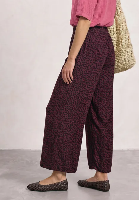 Mid Waist Straight Leg Hose mit Print rustic rouge Person trägt rosa Shirt, weite, dunkel gemusterte Hose, Netztasche und geflochtene Slipper vor hellem Hintergrund.