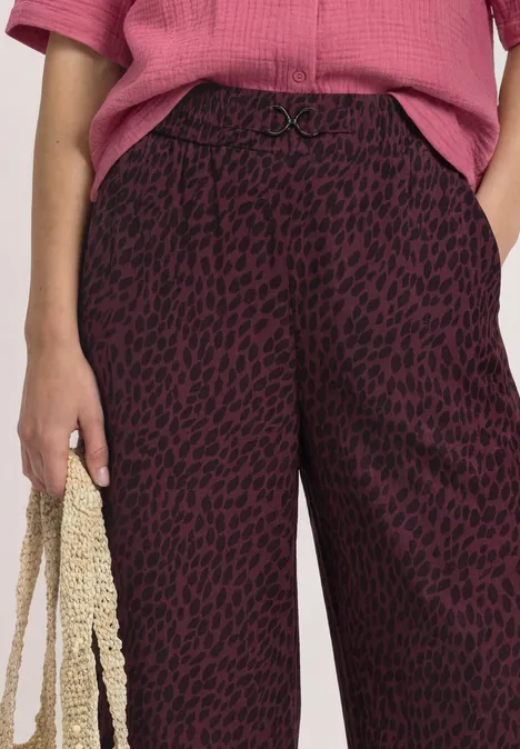 Mid Waist Straight Leg Hose mit Print rustic rouge Frau trägt weinrote Hose mit schwarzem Leoprint, rosafarbenes Oberteil und hält eine gehäkelte Tasche.