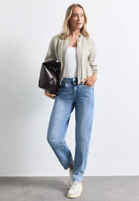 Jean taille haute à Barrel Leg, coupe ample Light Blue Random Washed Frau trägt weiße Sneaker, blaue Jeans, weißes Top und beige Jacke, hält schwarze Tasche.