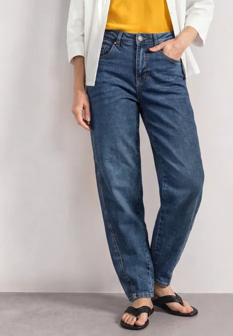 High Waist Barrel Leg Jeans im Loose Fit Medium Blue Washed Eine Person trägt blaue Jeans, gelbes Oberteil, weiße Jacke und schwarze Sandalen.
