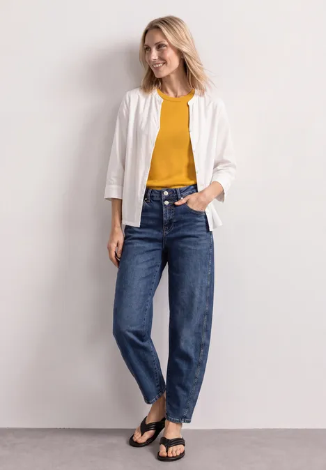 High Waist Barrel Leg Jeans im Loose Fit Medium Blue Washed Frau in weißer Jacke, gelbem Shirt, blauen Jeans und schwarzen Sandalen vor neutralem Hintergrund.