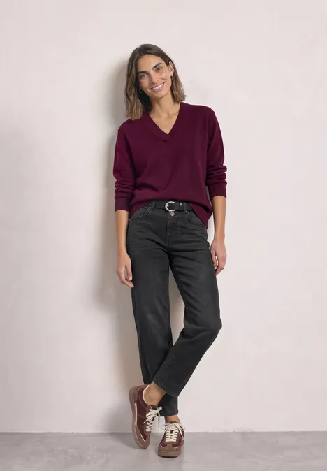High Waist Barrel Leg Jeans im Loose Fit Black Washed Frau in violettem Pullover, schwarzen Jeans und braunen Schuhen steht vor heller Wand.