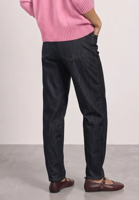 Person im Rückenansicht trägt dunkle Jeans, braune Loafers und einen rosa Pullover vor neutralem Hintergrund.