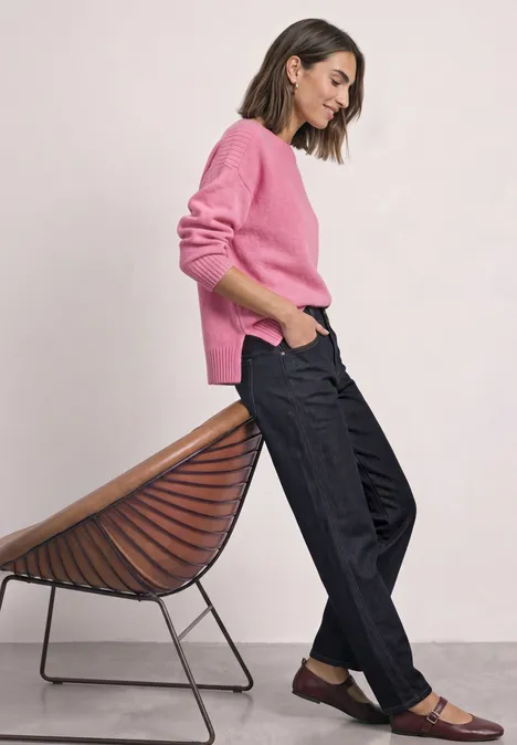 Frau in rosa Pullover und schwarzen Hosen lehnt lässig an einem modernen Stuhl.