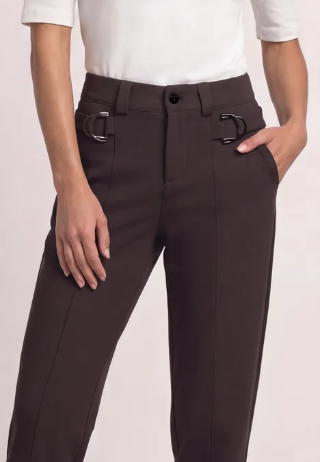Frau in dunkelbrauner Hose mit Gürtelringen und schmalem Schnitt, Hand in Tasche.