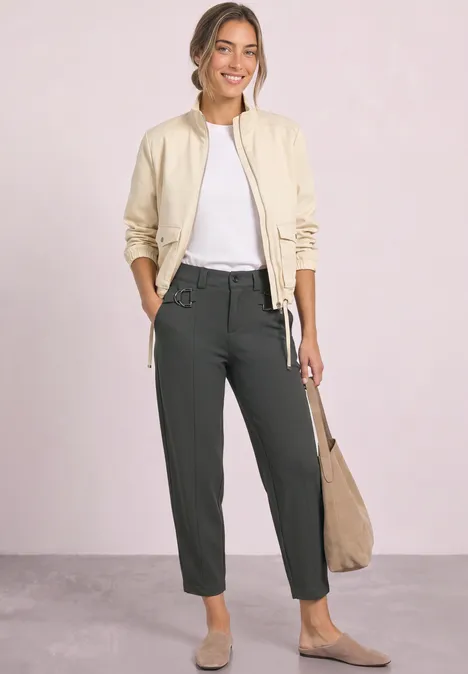 Frau mit heller Jacke, weißem Shirt und dunklen Hosen trägt Beutetasche und lächelt vor hellem Hintergrund.