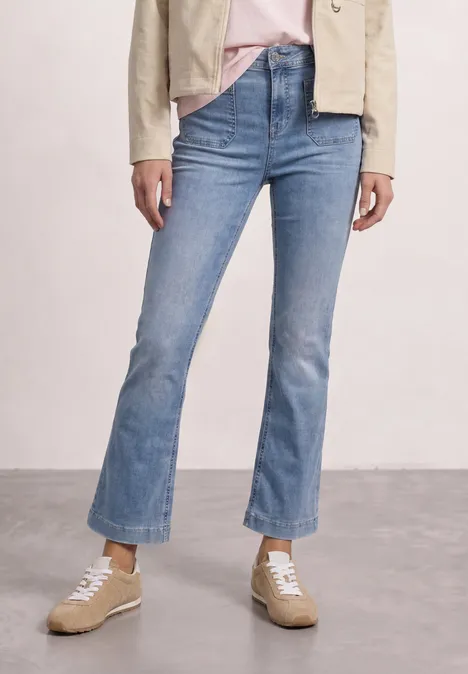 Frau trägt hellblaue Schlagjeans mit rohem Saum, beigen Sneakers, rosa Shirt und beiger Jacke.