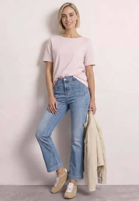 Junge Frau mit blonden Haaren trägt rosa T-Shirt, helle Jeans und Beige-Sneaker, hält eine Jacke in der Hand.