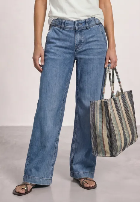 Jean taille moyenne à jambes larges, coupe ample mid blue washed Frau in weitem Jeans, weißem Oberteil und Sandalen hält gestreifte Tasche.