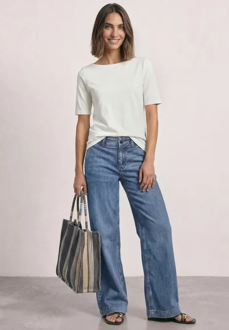 Jean taille moyenne à jambes larges, coupe ample mid blue washed Frau in weißem T-Shirt und weiten Jeans, mit gestreifter Tragetasche, lächelnd vor hellem Hintergrund.