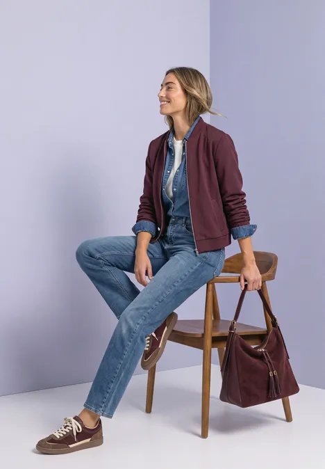 Jeans z prostymi nogawkami i guzikowymi detalami Medium Blue Random Washed Kobieta siedzi na krześle, ma na sobie dżinsy, koszulę i bordową kurtkę, obok leży torebka.