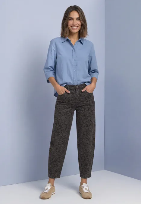 Jeans met High Waist en wijd uitlopende pijpen in een Loose Fit-pasvorm brown leo washed Een glimlachende vrouw in lichtblauwe blouse, bruine dessinbroek en sneakers voor een lichtblauwe achtergrond.
