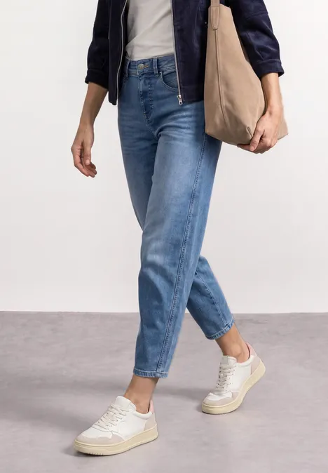 Person in blauen Jeans, weißen Sneakern und dunkelblauer Jacke trägt lässig eine Tasche über der Schulter.