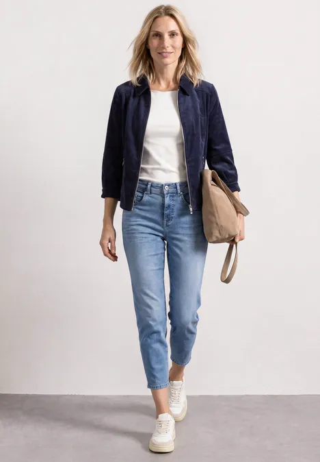 Eine Frau in weißem Shirt, dunkelblauem Blazer und Jeans trägt eine Tasche und geht lächelnd.