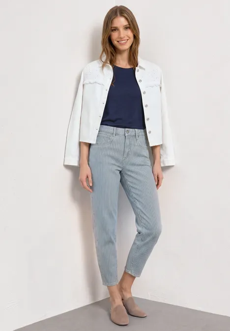 Vrouw draagt witte spijkerjas over donkerblauw shirt en lichtgrijze broek, staand voor lichte achtergrond.