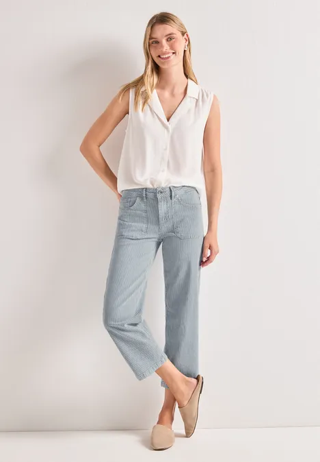Vrouw in mouwloze witte blouse, lichtblauwe jeans en beige loafers voor lichte achtergrond.