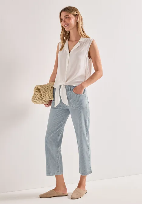 Vrouw in wit geknoopt hemd, lichtblauwe jeans, beige loafers en stro handtas voor witte muur.