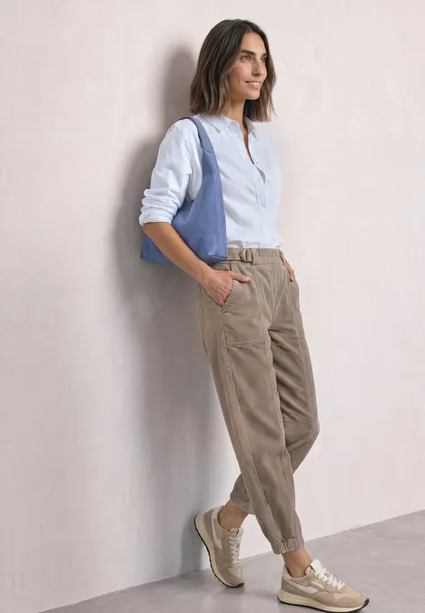 Une femme appuyée contre un mur, chemisier blanc, pantalon beige en velours côtelé, gilet bleu sur l’épaule et baskets.