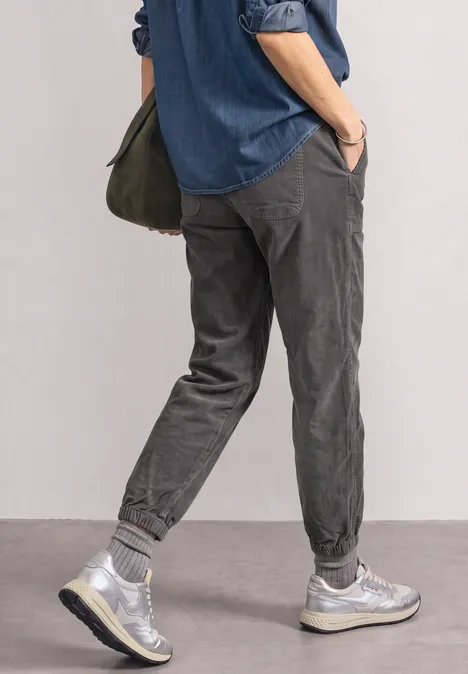 Person in knöchellangen, grauen Chino-Jogginghosen, Sneakers, Jeanshemd und grüner Umhängetasche, seitlich stehend.