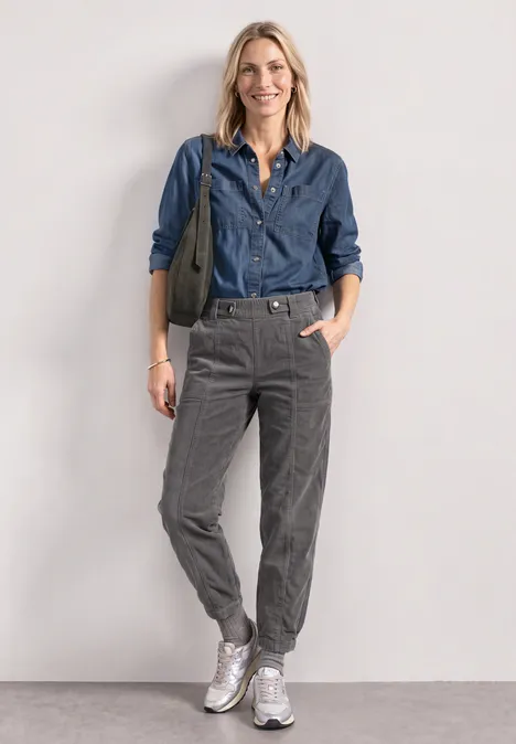 Eine lächelnde Frau trägt eine Jeansbluse, graue Hose, weiße Sneaker und hält eine grüne Tasche.