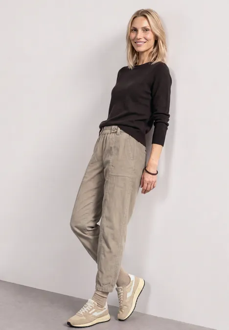 Eine lächelnde Frau vor weißem Hintergrund, in schwarzem Pullover, beigen Hosen und hellen Sneakers.