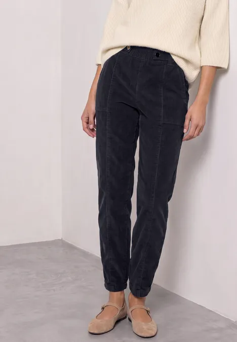 Schwarze, taillierte Cordhose mit schmalem Beinabschluss, kombiniert mit hellem Oberteil und Sandalen.