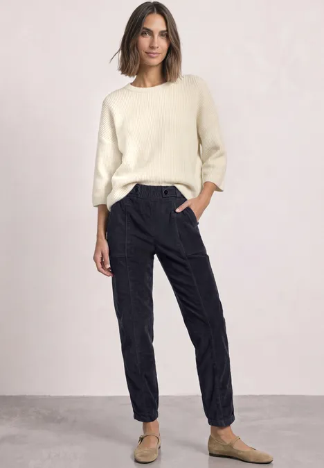 Eine Frau steht in einem hellen Pullover, dunklen Hosen und braunen Hausschuhen vor einer neutralen Wand.