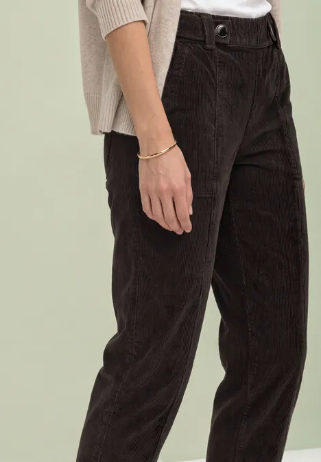 Teilansicht einer Person in dunkler Cordhose, hellem Oberteil und sichtbarem Armband.