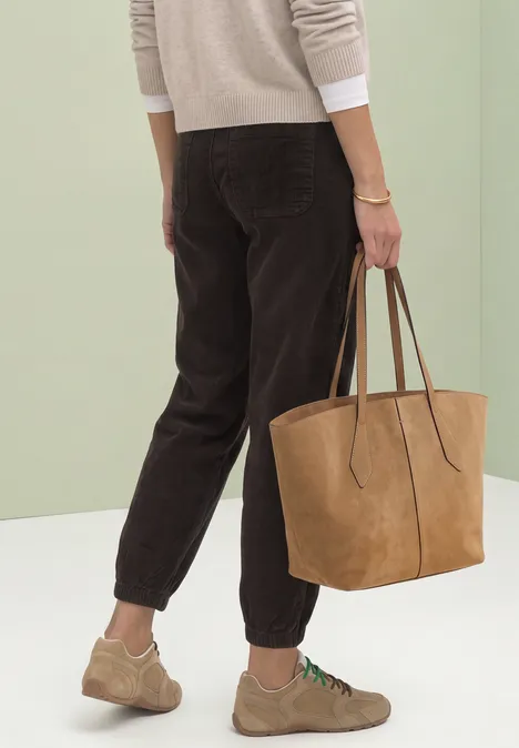 Frau mit beigem Pullover, brauner Cordhose, hellen Sneakern und großem Wildleder-Shopper.