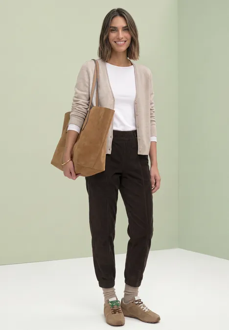 Eine Frau mit heller Strickjacke, weißem Shirt, dunkler Hose, braunen Boots und Beuteltasche vor neutralem Hintergrund.