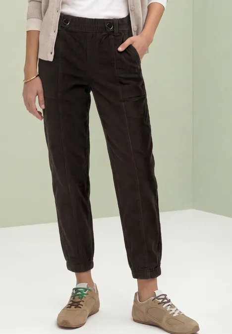 Person trägt schwarze Cordhose mit Gummibund und beige Sneaker vor hellgrünem Hintergrund.