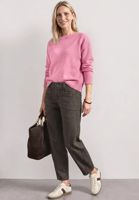 Vrouw in roze trui, donkeren broek, witte sneakers en bruine tas, ontspannen pose voor lichte achtergrond.