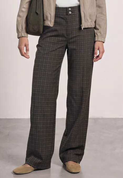 Wijdvallende donkergrijze broek met subtiel ruitpatroon, gecombineerd met een lichte jas en beige loafers.