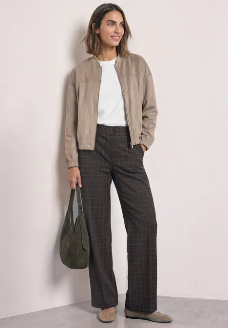 Vrouw in beige suède bomberjas, wit shirt en wijde geruite broek met groene tas voor lichte achtergrond.