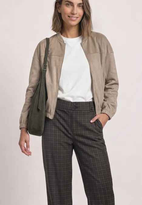Vrouw in beige jas, wit t-shirt, geruite broek en schoudertas tegen lichte achtergrond.