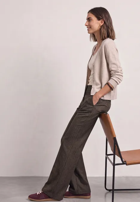 Pantalon taille haute Straight Leg avec détail boutonné black coffee Une femme assise de profil sur un tabouret, vêtue d'un cardigan clair, d'un top blanc et d'un pantalon ample marron.