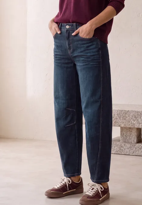 High Waist Barrel Leg Jeans im Loose Fit Dark Blue Washed Person steht vor hellem Hintergrund in dunkelblauen Jeans, weinrotem Pullover und Sneakern.