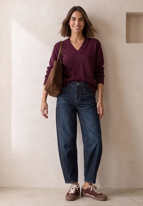 High Waist Barrel Leg Jeans im Loose Fit Dark Blue Washed Eine Frau mit weinrotem Pullover, blauen Jeans und brauner Tasche steht lächelnd an einer Wand.