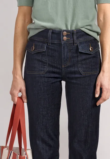 Frau mit grünem Shirt, dunklen High-Waist-Jeans mit Taschen, hält eine rot-orange Handtasche.