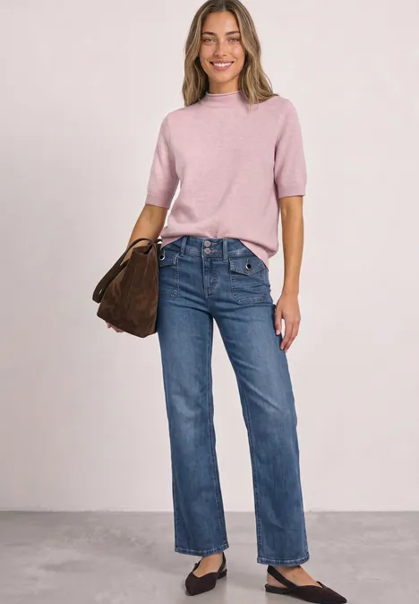 Mid Waist Straight Leg Jeans im Casual Fit Mid Blue Washed Frau trägt rosa Kurzarm-Pullover, blaue Jeans, schwarze Ballerinas und braune Tasche.