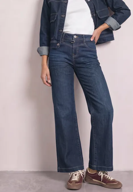 Une personne porte un jean flare bleu foncé taille haute, un t-shirt blanc, une veste en jean et des baskets marron.