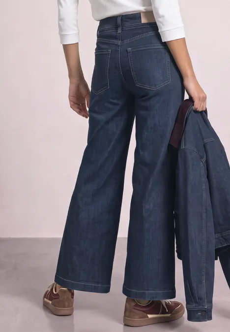 Jean taille haute, coupe large et jambe évasée, vue de dos, main tenant une veste en jean assortie.