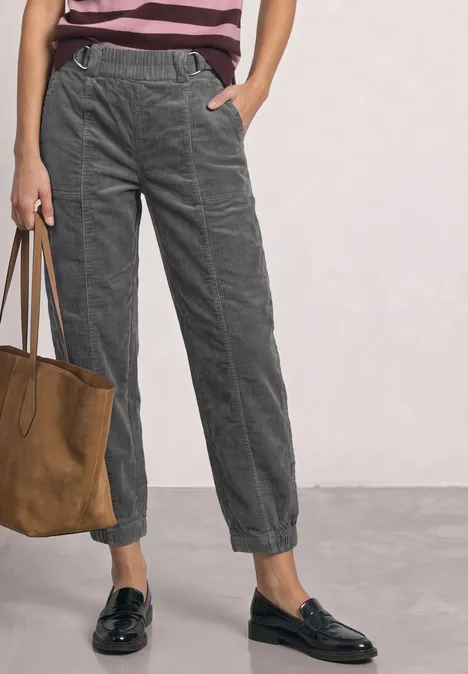 High Waist Barrel Leg Hose mit Elastikbund eclipse grey Frau stehend mit grauen Cord-Joggerhosen, rosa Streifenshirt, schwarzen Loafers und brauner Wildledertasche