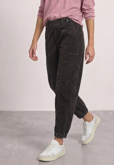 Dunkelbraune Cordhose mit lockerer Passform, hochgekrempelt, kombiniert mit pinkem Oberteil und weißen Sneakern.