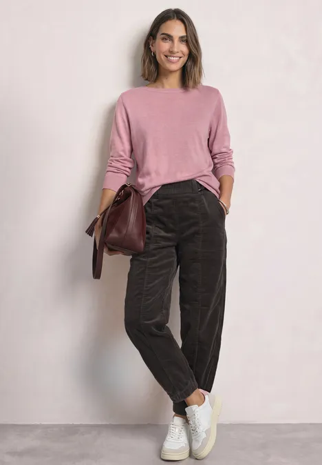 Eine Frau trägt einen rosafarbenen Pullover, braune Cordhosen, weiße Sneaker und hält eine Tasche.